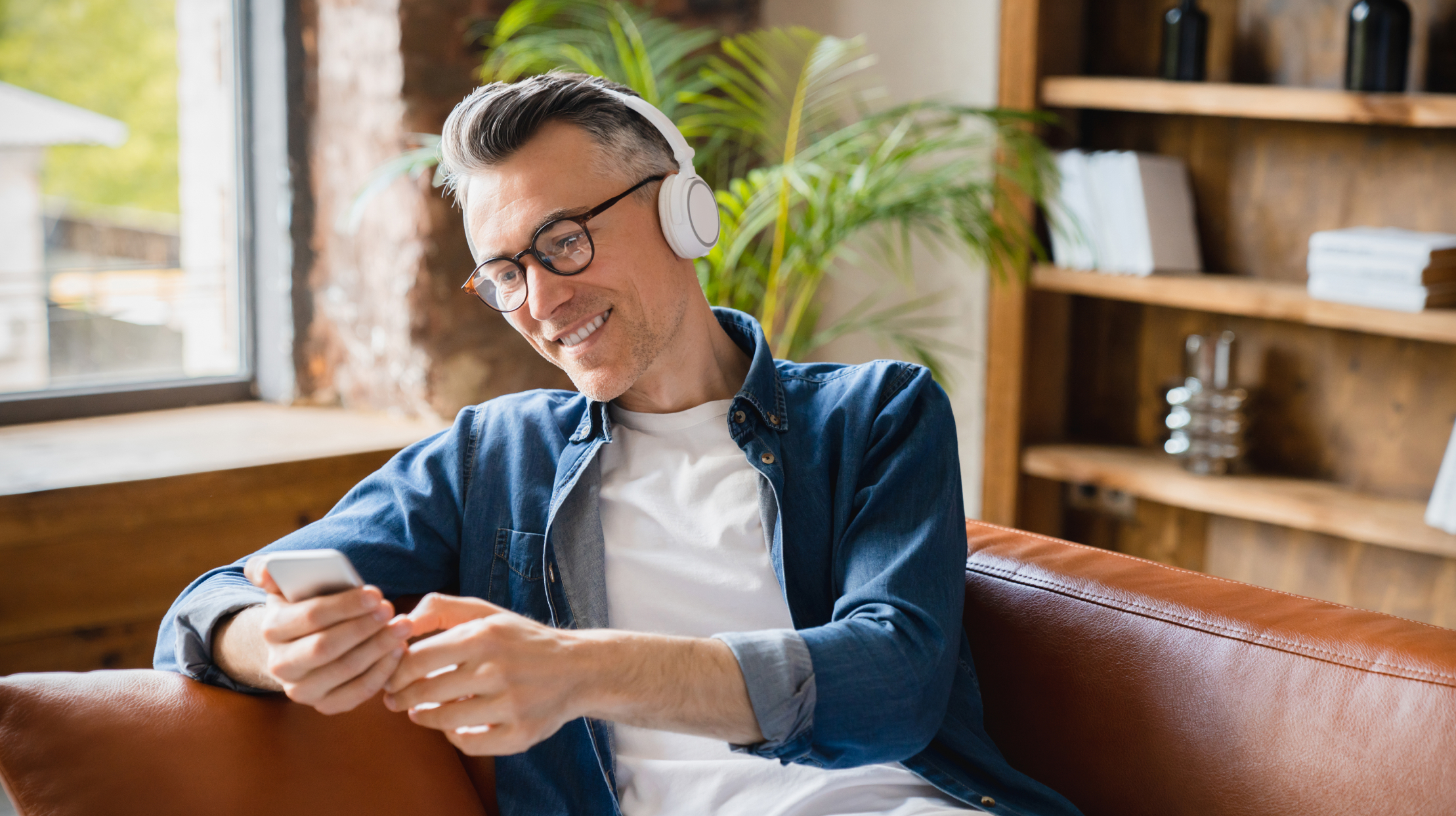 Smiling man wearing headphones sitting in a sofa using a smartphone in a brightly lit office room.