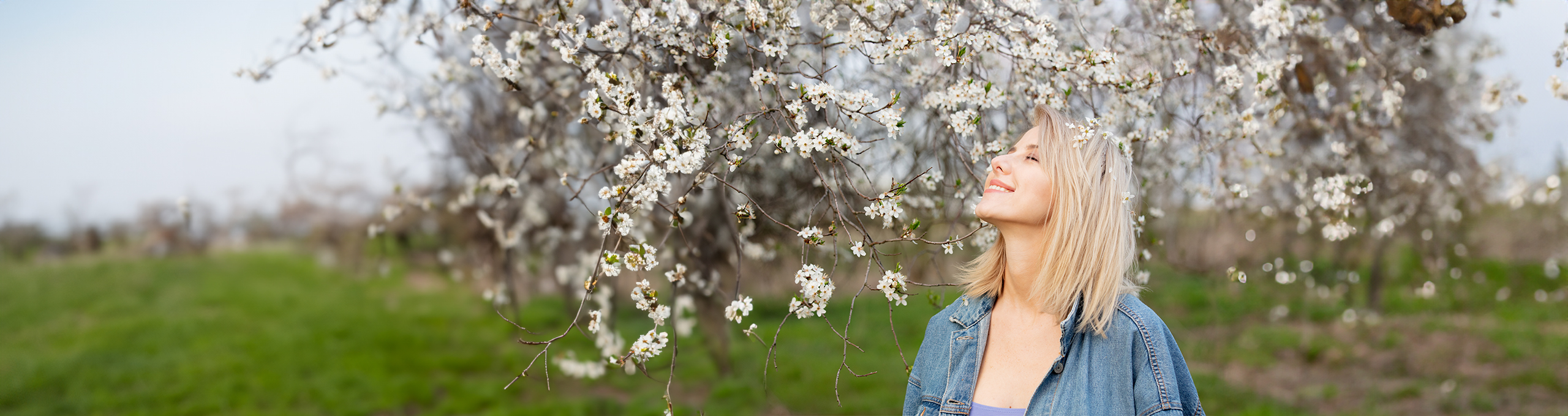 Glad kvinna står under ett blomstrande körsbärsträd i solen med slutna ögon.